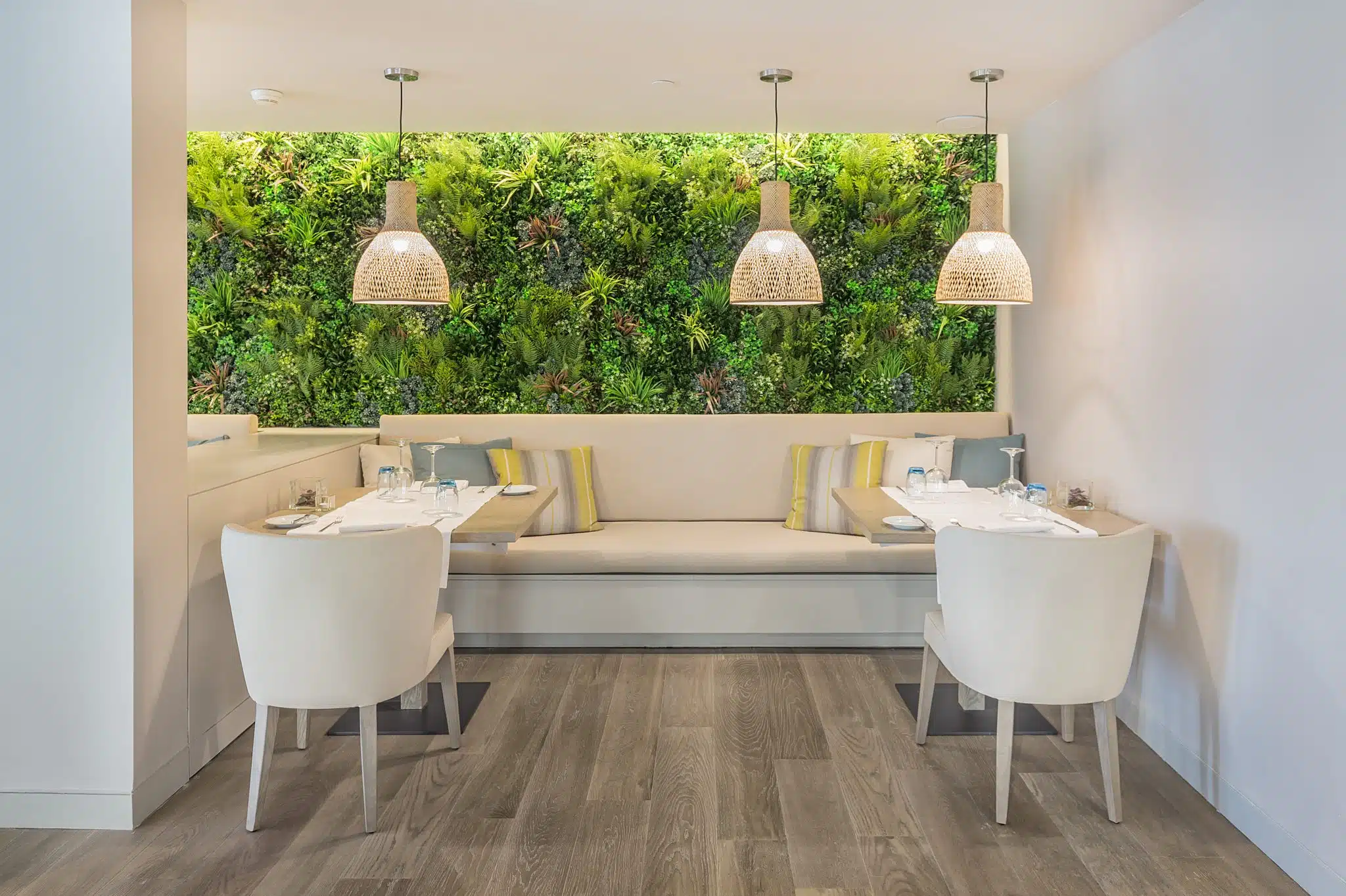 A modern restaurant interior with two tables and white chairs. A cushioned bench runs along a wall adorned with a vertical garden. Three woven pendant lights hang above, and light wood flooring adds warmth to the space.