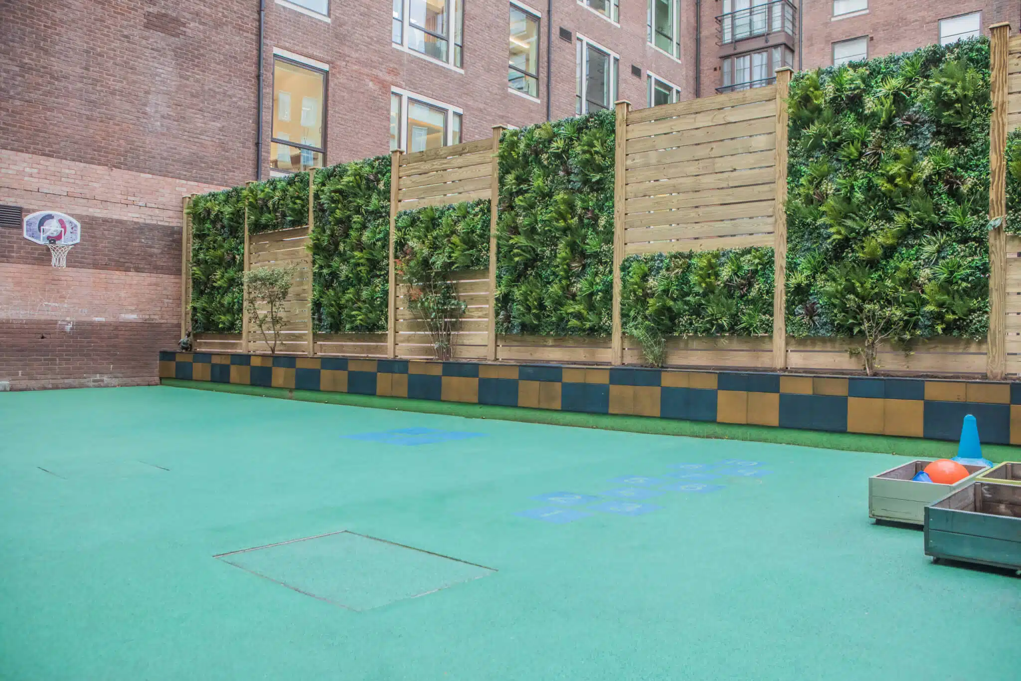 A school playground with a green surface, featuring a wall covered with greenery. Wooden panels support the plants. On the left, a basketball hoop is mounted on a brick wall. On the right, there are storage bins with colorful balls.