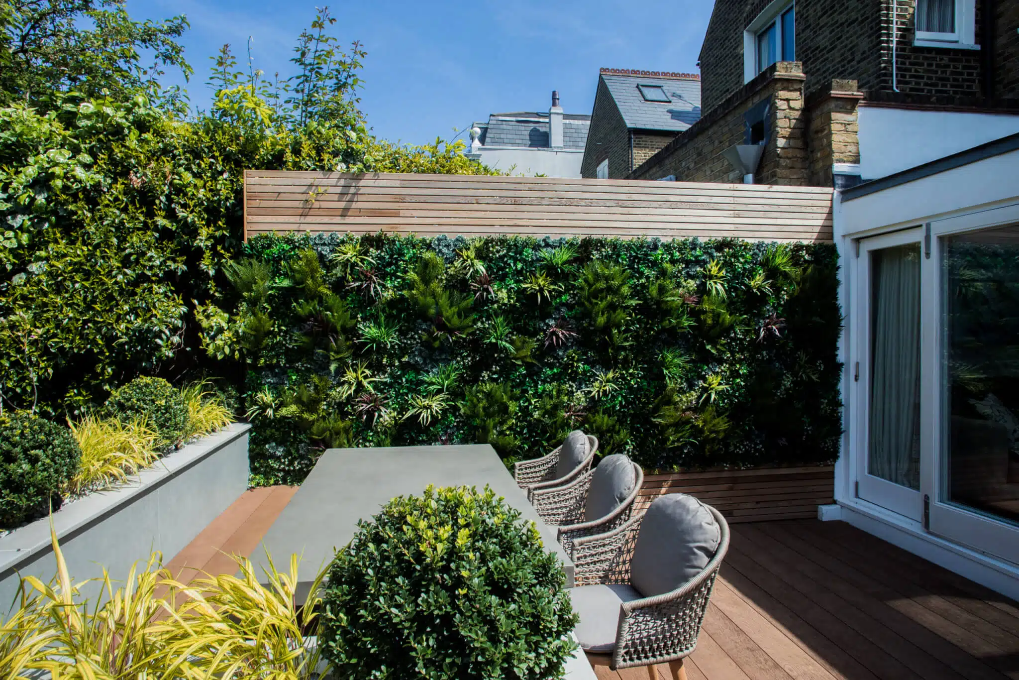 A modern outdoor patio with a large dining table surrounded by wicker chairs. Theres a lush green vertical garden on the wall, wooden fencing, and potted plants. The area is adjacent to a white building with large glass doors.