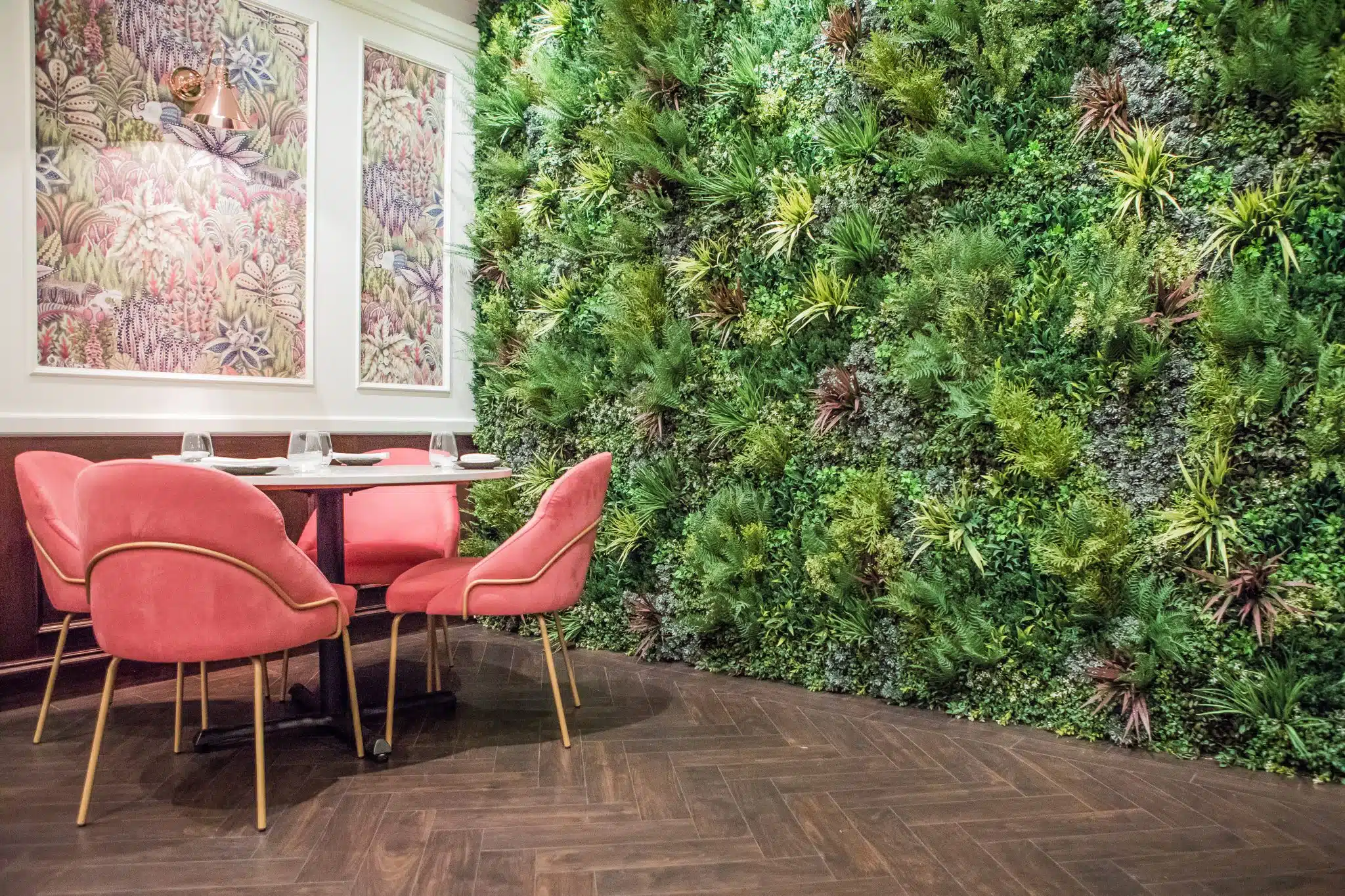 A cozy dining area with a round table, set for four, surrounded by plush pink chairs. The interior features a lush green vertical garden on one wall and framed botanical artwork on another. The flooring is dark wood with a herringbone pattern.