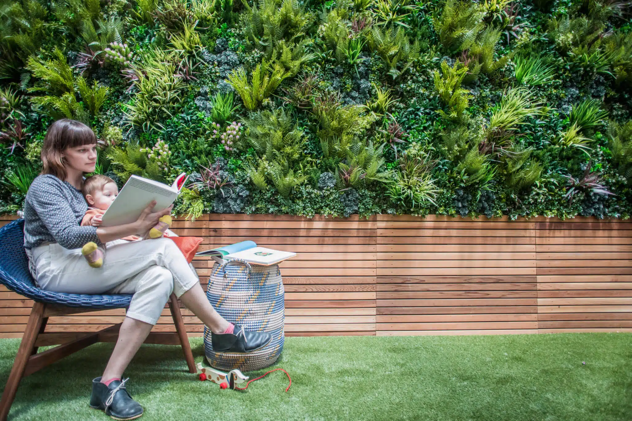 A woman sits in a chair reading a book, holding a baby on her lap. They are in a garden-like space with a wooden wall and lush green plants. A small table with books and a blanket is beside them, on a grassy surface.