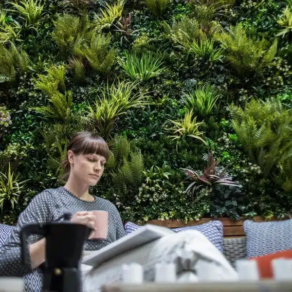 A woman sits on a couch, holding a pink mug and a notebook, in front of lush green wall foliage panels. These vibrant installations are filled with various plants, creating an enchanting backdrop. A coffee pot is visible on the table in the foreground.