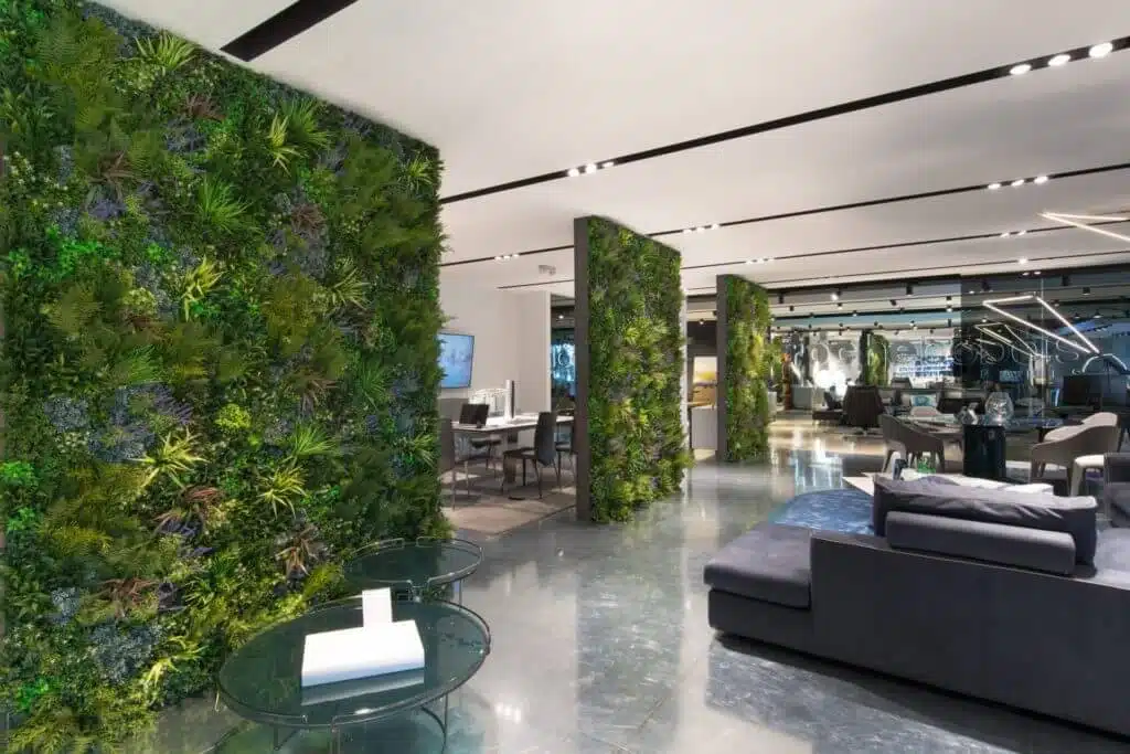 Modern office space with large living plant walls reminiscent of a Roche Bobois Showroom Green Wall in Hollywood. It boasts polished concrete floors and sleek furniture, including glass tables and a dark couch, crafting an open and green ambiance that feels straight out of California.