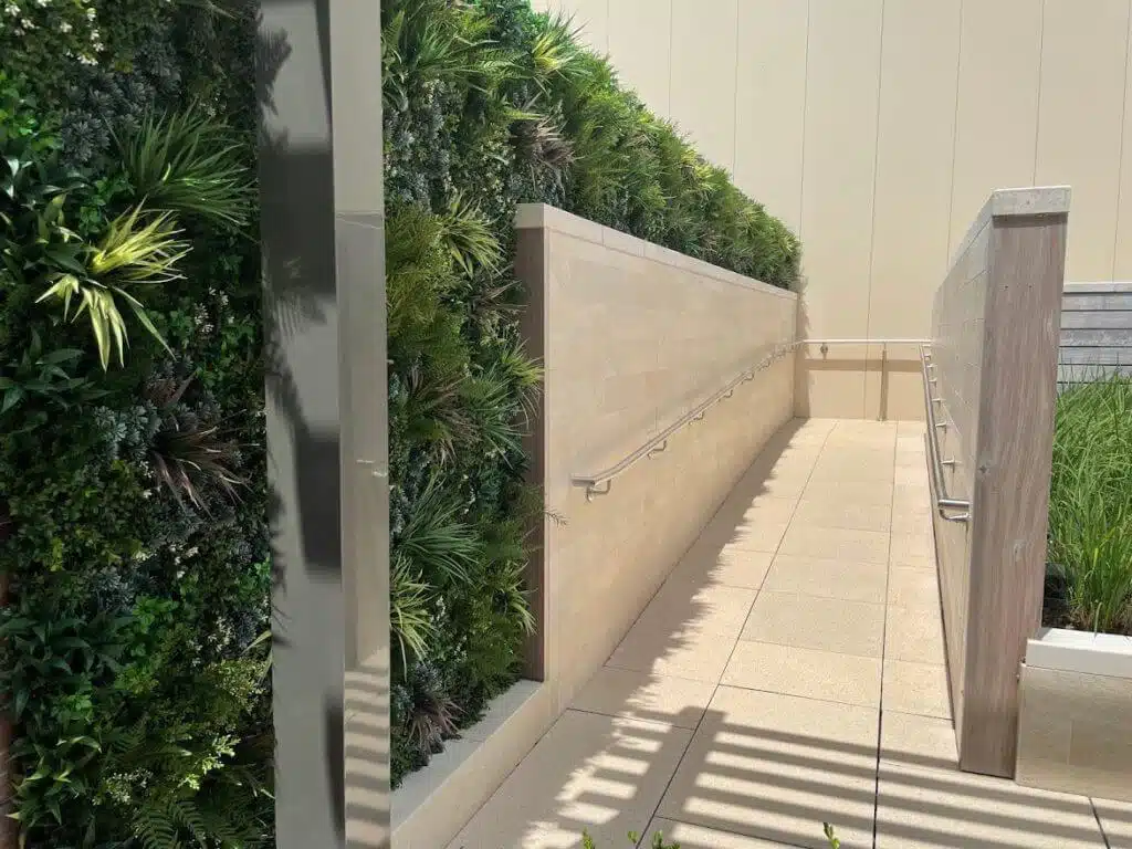 A modern outdoor walkway with a beige tile path and safety railings on both sides leads to the fitness center. To the left, a lush green wall adds vibrancy, while on the right, a wooden and stone wall features plants at its base. Natural light casts gentle shadows in Houston, Texas.