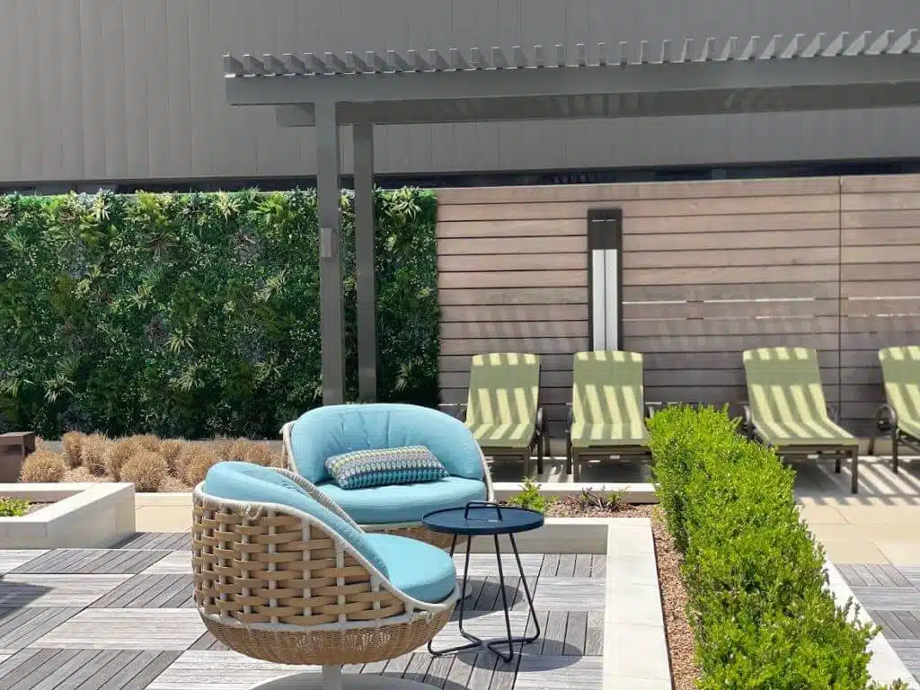 Modern outdoor patio with woven chairs and blue cushions under a pergola, resembling a serene oasis. A lush green wall enhances the space next to wood fencing. Striped lounge chairs line the wall beside manicured bushes, reminiscent of an upscale fitness center in Houston, Texas.