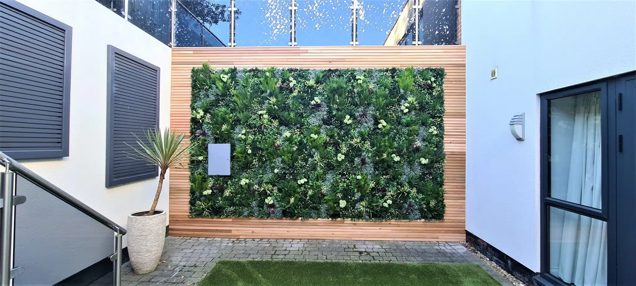 A courtyard exudes luxury with a green wall of Luxury Boxwood Panels, towering glass railings, and a potted plant on the left. Gray shutters contrast white walls, while artificial grass softens the stone-paved ground.
