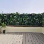 A spacious rooftop terrace features a wooden floor and is enclosed by a low yellow wall. Lush artificial greenery with purple flowers lines the perimeter. Terracotta pots with plants are placed to one side, and the sky is clear above.