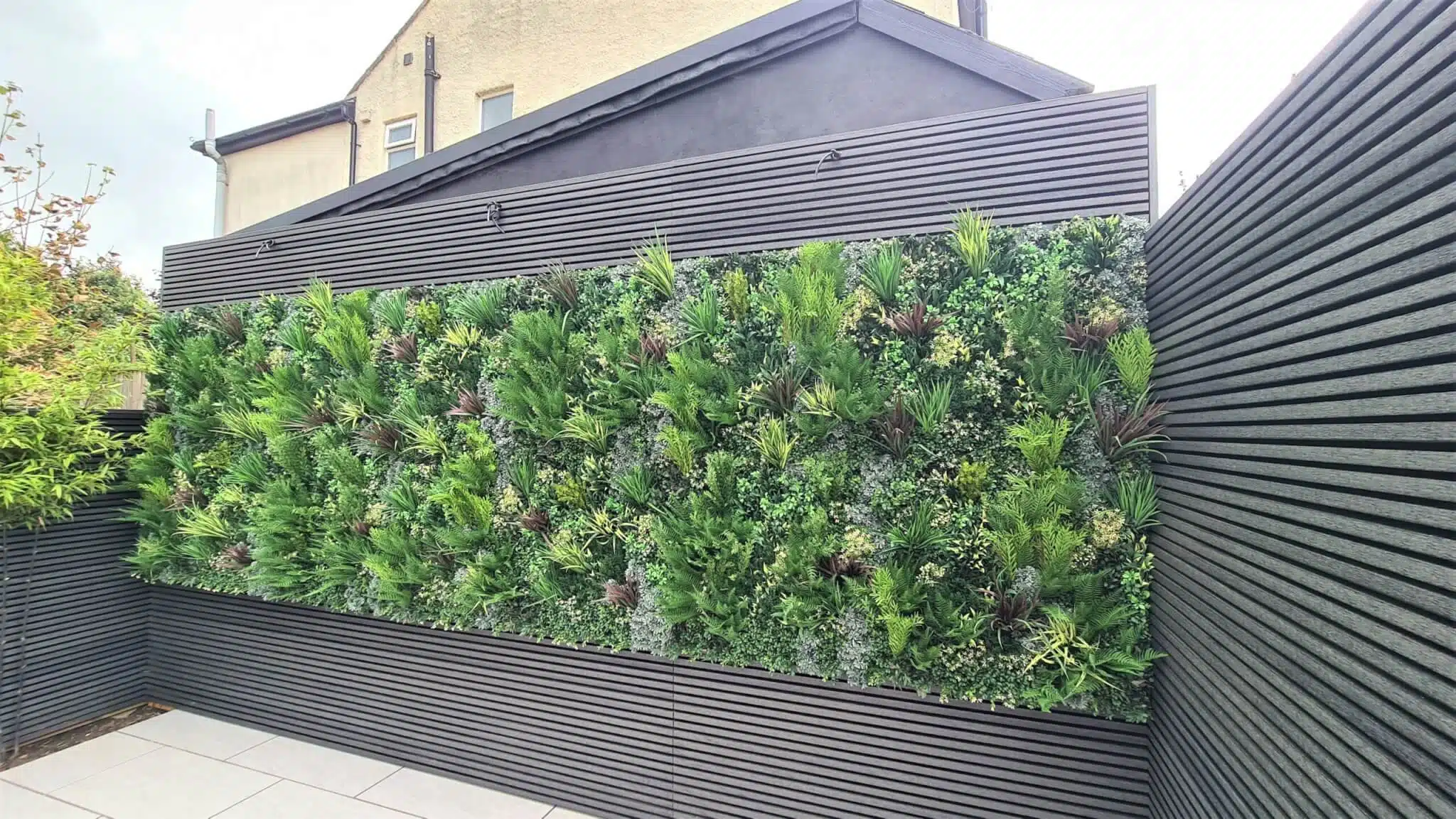 A vertical garden with lush green plants is mounted on a black slatted wall in a backyard. The modern setup stands against a dark building, creating a striking contrast. The flooring is light-colored tile.
