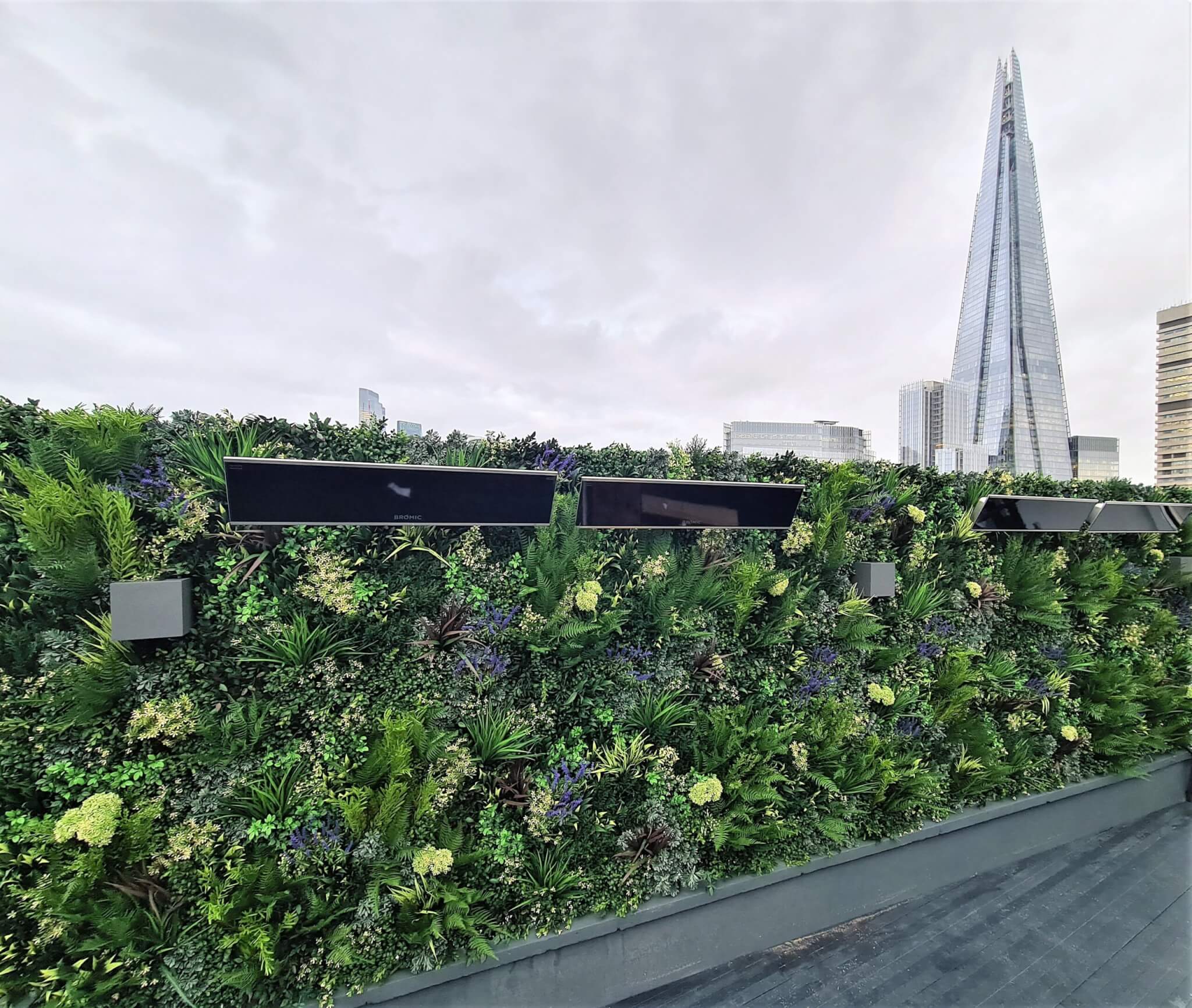 A lush green wall with various plants, featuring black rectangular panels. In the background, the tall, pointed structure of The Shard is visible against a cloudy sky.