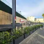A modern outdoor restaurant adorned with a ZIZIGO sign features luxury boxwood panels, a paved walkway, and wooden and glass railings separating it from an elevated area, all under a cloudy blue sky backdrop.