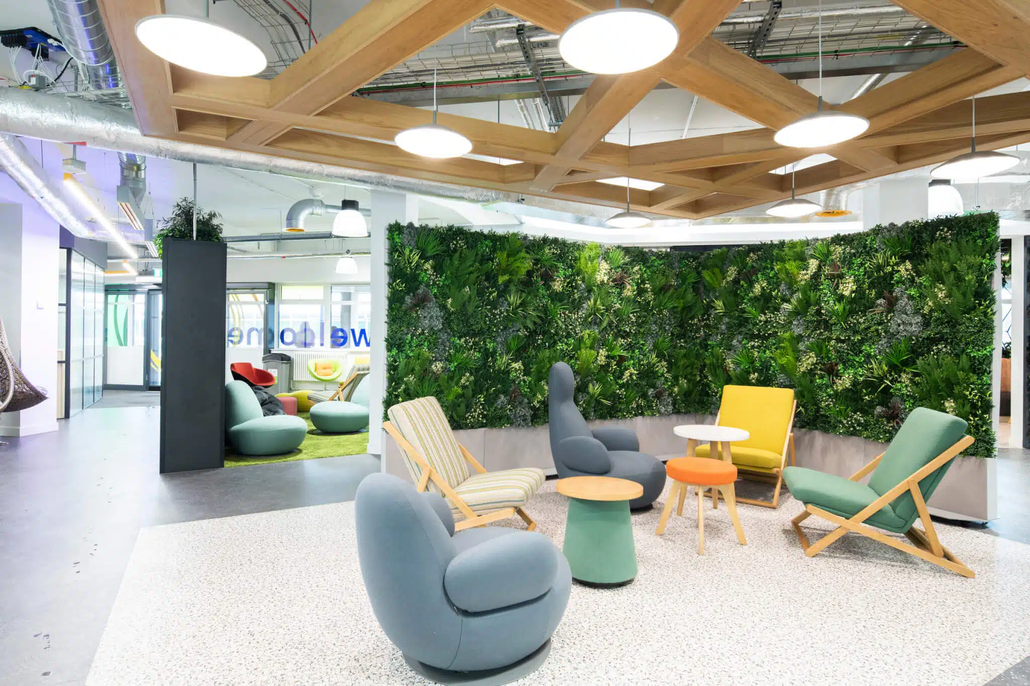 A modern office lounge features colorful chairs and small round tables on a speckled floor. A lush green plant wall, enhanced by Modular Green Wall Partitions, serves as the backdrop. The ceiling showcases a wooden grid design with circular lights and exposed pipes and ducts.