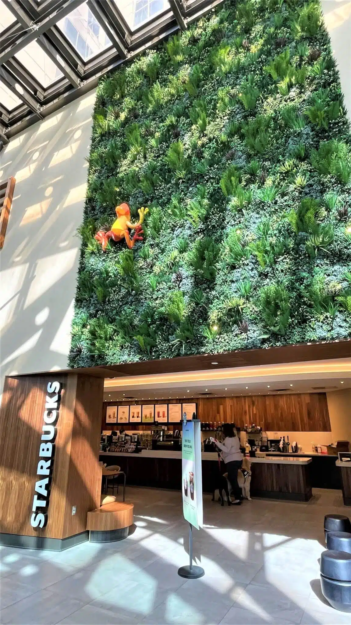 A tall, lush artificial green wall dominates a bright interior space in Dallas, with sunlight streaming through a glass ceiling. A small figure of the Pink Panther is climbing the wall. Below, a Starbucks counter and sign are visible with people ordering amidst the bustling Texas vibe.