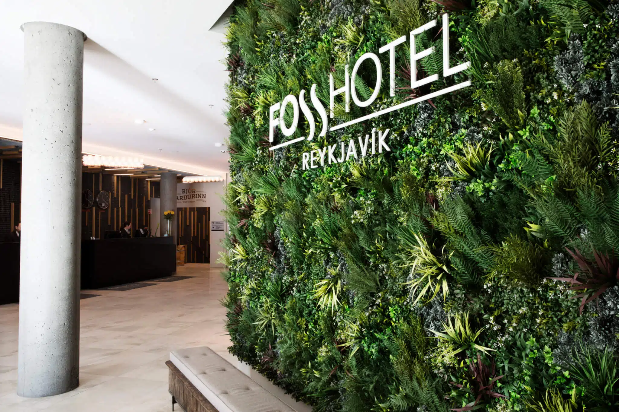 Hotel lobby with a lush green vertical garden displaying the name Fosshótel Reykjavík. The reception desk is visible in the background, with modern, minimalist decor and soft lighting.
