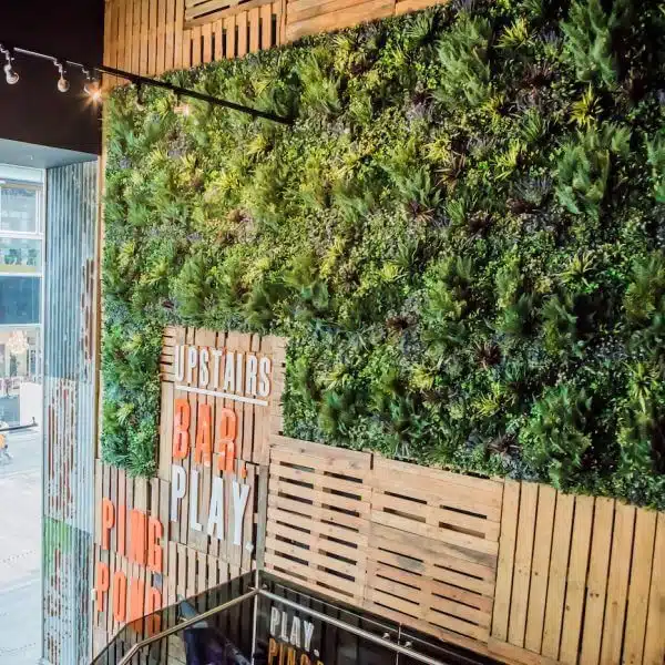 In a chic London setting, this green vertical garden wall with wooden slats adds elegance indoors. Featuring signs for UPSTAIRS in white and BAR PLAY in orange, the stairs lead down to large windows filling the space with natural light at Westfield Shopping Centre's PROJECT SPOTLIGHT: Bat & Ball.