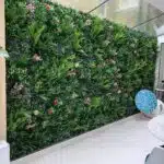 The Spring Pink Colour Box features a vertical indoor garden with lush greenery and spring pink flowers brightening the wall. A light wood-paneled section complements the blue-patterned chair and white marble table, while natural light from a large window enhances this vibrant space.