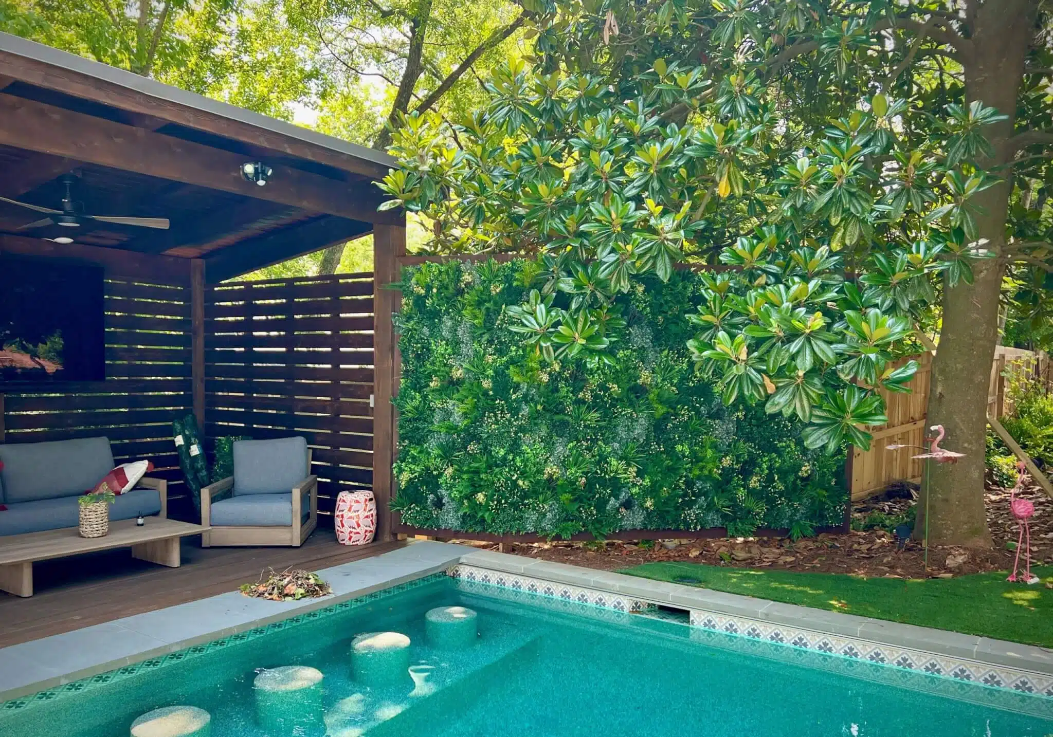 A backyard pool area with clear blue water and stepping stones. To the left, theres a covered wooden seating area with gray chairs and decorative pillows. Lush green foliage surrounds the pool, and a pink flamingo decoration is in the grassy area.