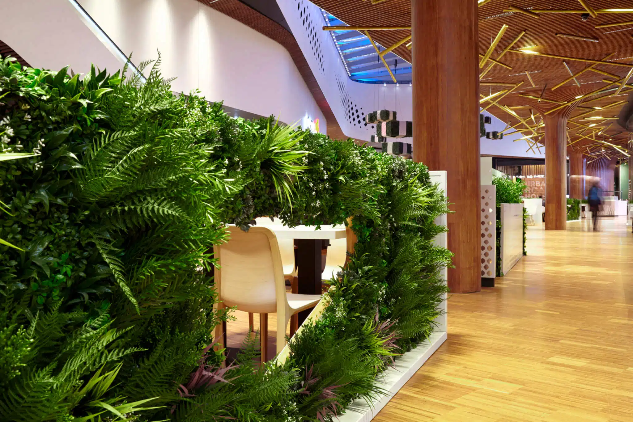 A modern indoor space with wooden flooring and ceiling, featuring a dining area separated by lush green plant partitions. Curved walls and ceiling lights add to the contemporary design.