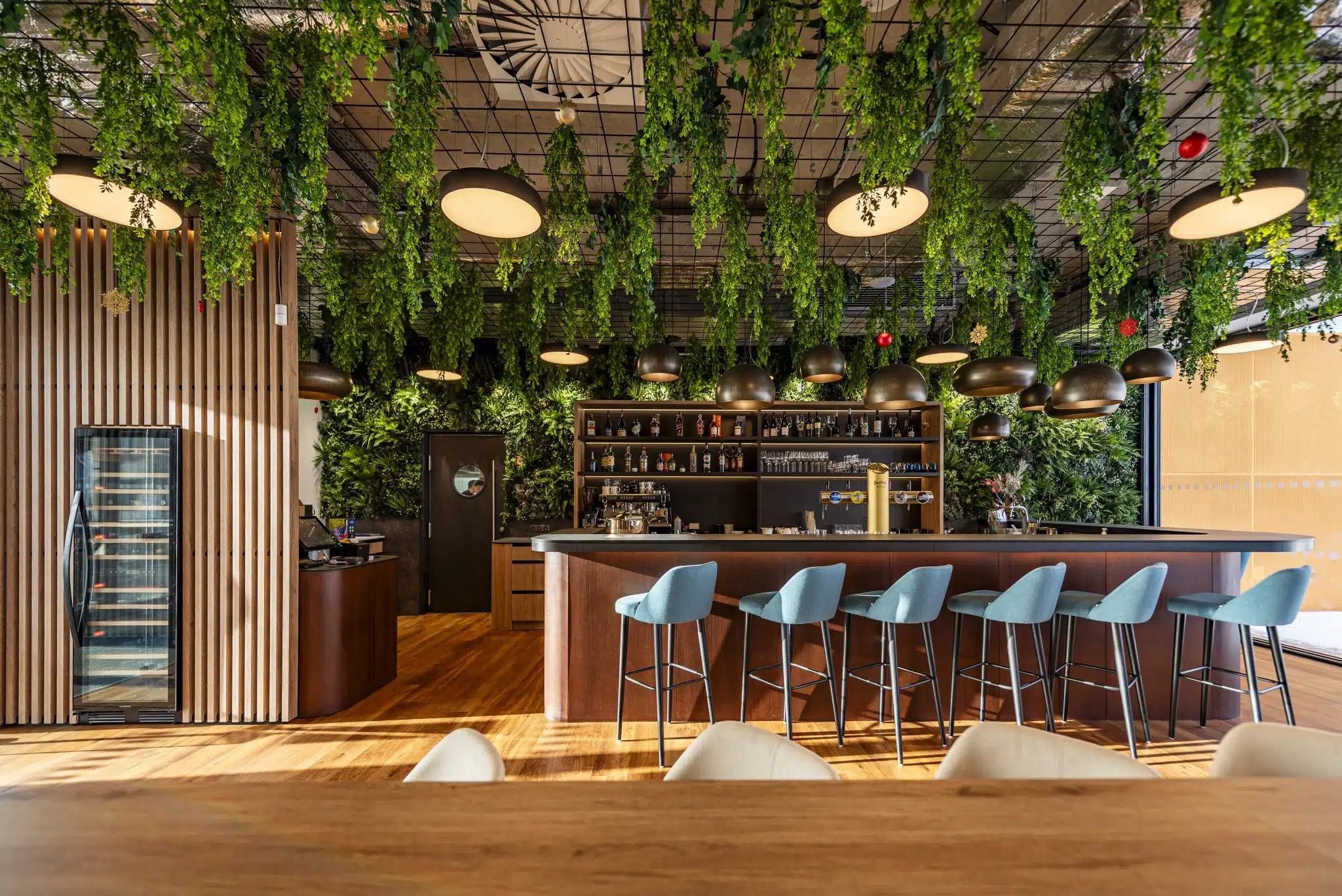 Bar interior featuring a long counter with blue stools, surrounded by hanging green plants. Wooden accents and shelves with bottles are visible. Warm lighting creates a cozy atmosphere, with large pendant lights and a modern, stylish design.