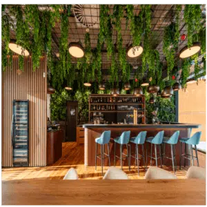 The bar interior features a wooden counter with high blue stools, while the ceiling and walls are embellished with artificial plants, foliage, and greenery. Brass light fixtures hang above the bar, shelves are stocked with bottles, and there's a glass fridge on the left. The look is completed by hardwood floors.
