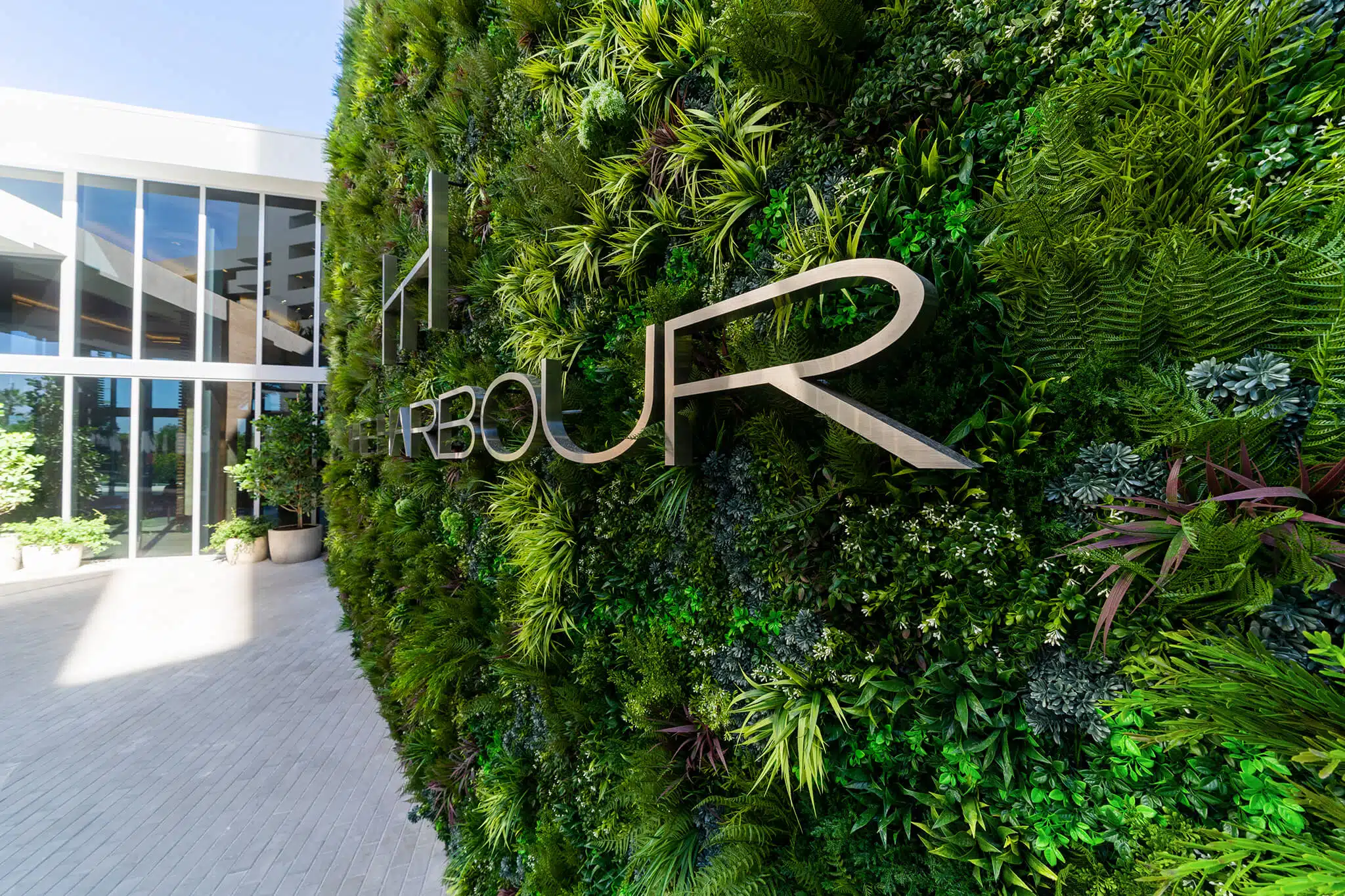 A vertical garden wall features a mix of lush artificial plants from the Artificial Plants & Flowers collection, paired with metallic letters spelling HARBOUR. To the left, a modern glass building entrance with planters is bathed in bright, natural light.
