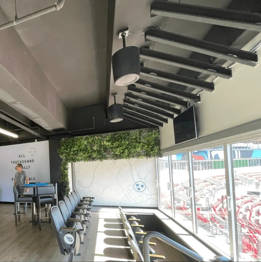 A modern stadium suite with a row of high chairs facing large windows overlooking the field. The suite features a bar area, green wall decor, and a flat-screen TV. Large text on the wall reads ALL TOUCHDOWNS BIG OR SMALL, THEY SCORE EQUALLY.