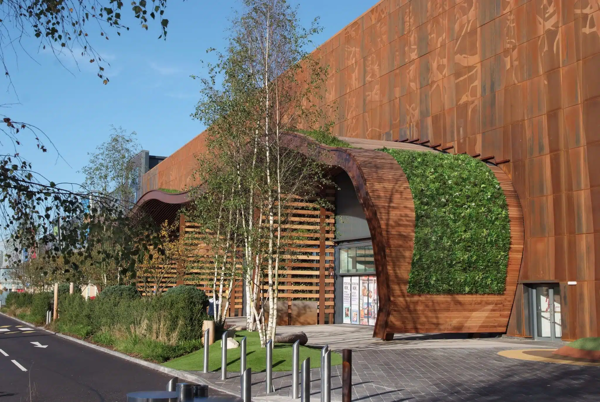Modern building with rust-colored, wavy metal facade and green wall. Birch trees and shrubs line the entrance. Sunlit exterior with a clear blue sky. Bike racks and a paved walkway are in the foreground.