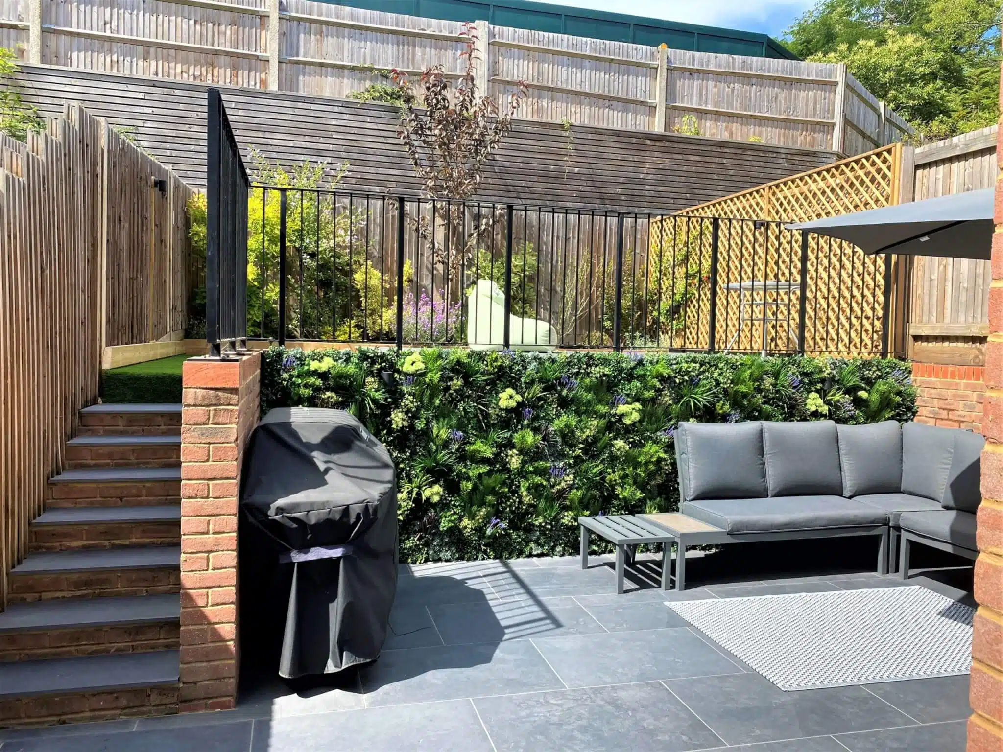 A modern backyard features a seating area with a gray outdoor couch and table on slate tiles. A green wall with plants is behind the couch. A barbecue grill with a cover is beside steps leading to a raised garden area with more greenery.