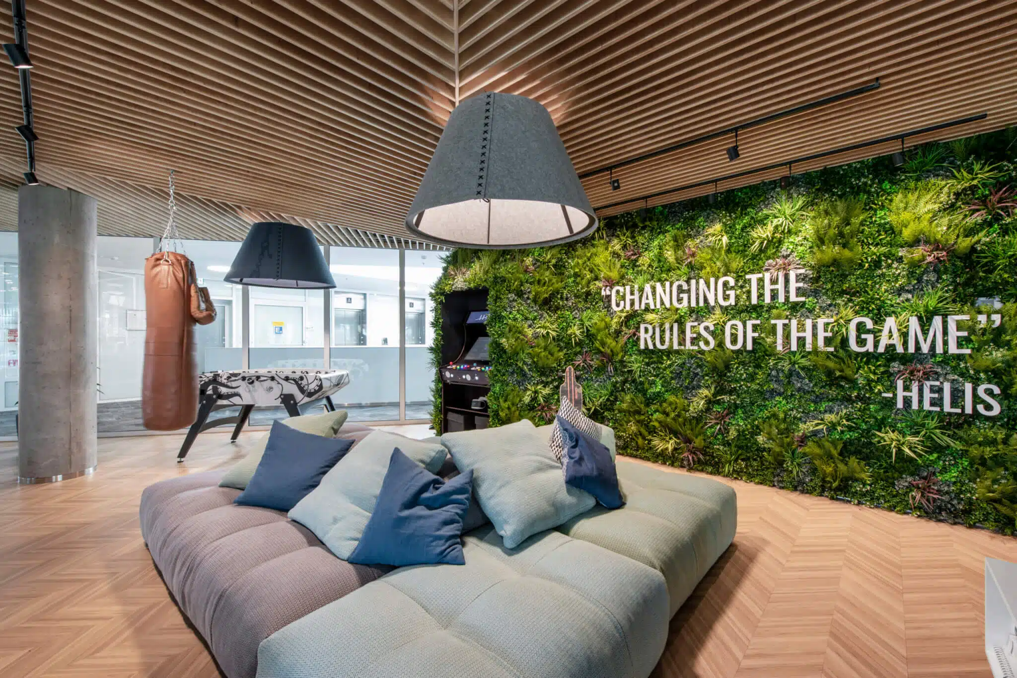 Modern lounge with a cushioned sectional sofa and pillows, a punching bag, foosball table, and arcade machine. A wall displays a greenery backdrop with the words CHANGING THE RULES OF THE GAME attributed to Helis.