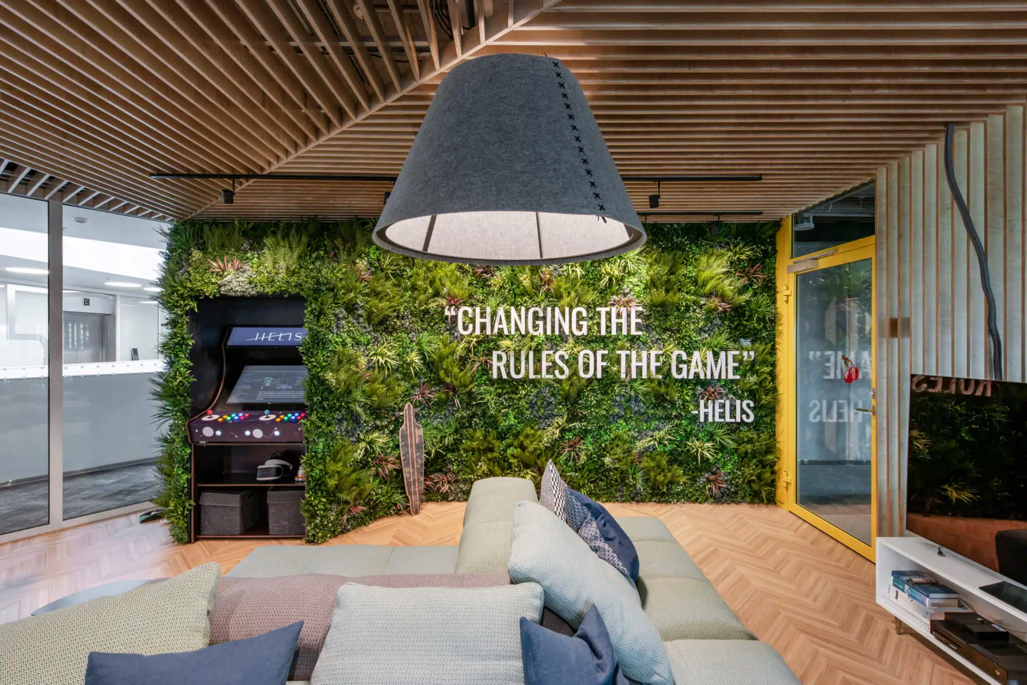 Modern office lounge with green plant wall displaying Changing the Rules of the Game and Hello. An arcade machine is on the left. A large, grey lampshade hangs above a light-colored sectional sofa with multiple cushions.