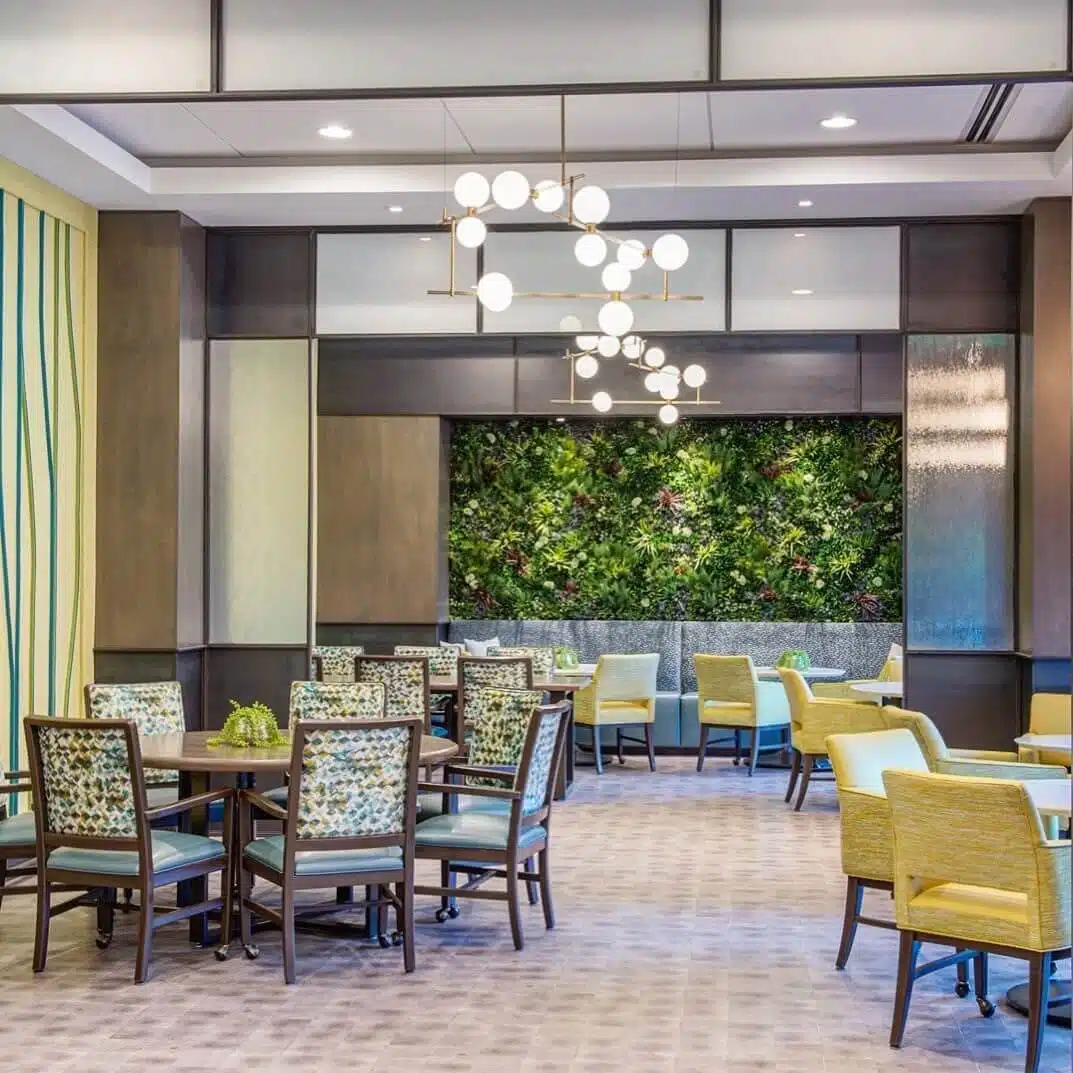 A modern dining area with wooden tables and chairs featuring patterned cushions. A lush green vertical garden is on the back wall. Contemporary spherical light fixtures hang from the ceiling. Neutral tones with green accents dominate the space.