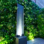 A vertical water feature with subtle illumination stands out against a lush green wall. The leafy backdrop creates a serene ambiance, enhanced by the Soft Lavender Colour Box. A wooden deck and part of a brick wall complete the setting.
