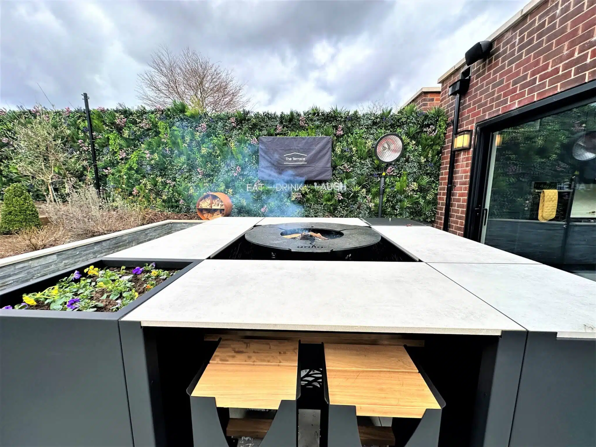 Outdoor kitchen setup with a central grill, surrounded by a stone counter. Smoke is rising from the grill. Colorful flowers are planted on the left side. A green hedge and a brick wall form the backdrop.