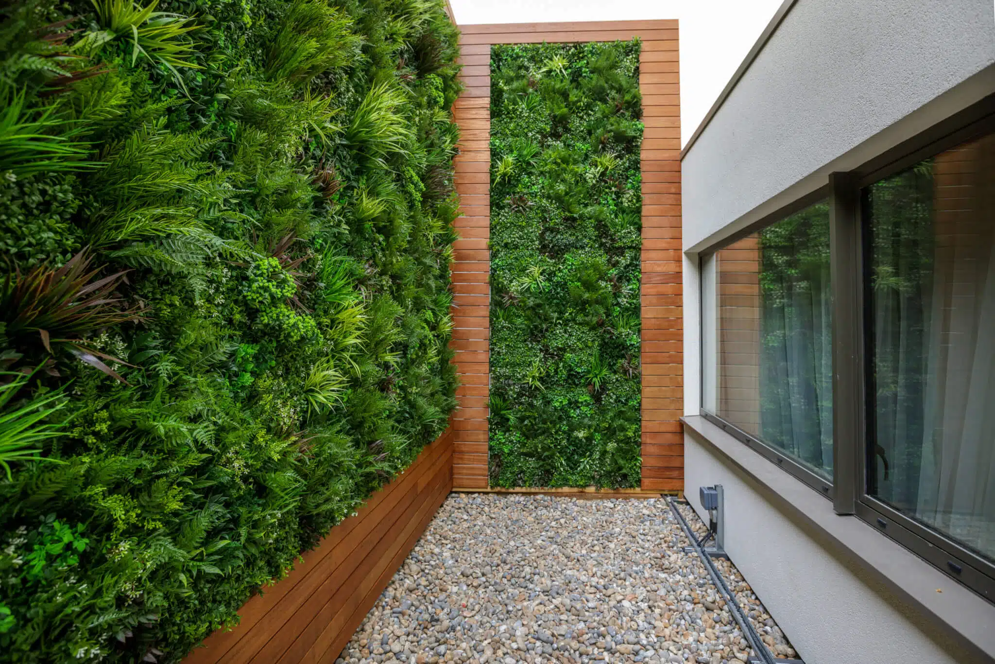 A modern courtyard features vertical green walls filled with lush plants, bordered by wooden paneling. A window on the right reflects the greenery. The ground is covered with small pebbles, creating a minimalist design.
