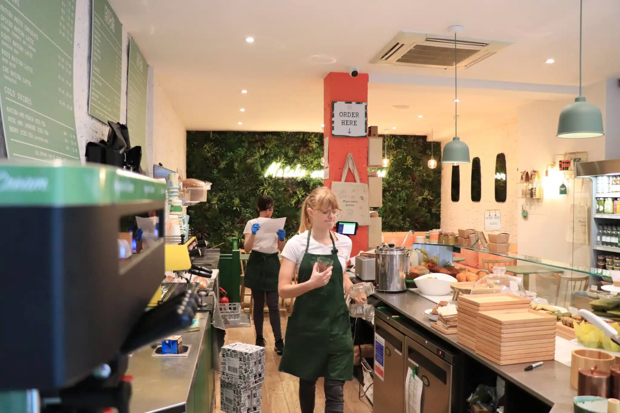 A bustling sandwich shop with two employees in green aprons. One is in the foreground holding a small bag, walking near a counter with food prep items. The interior is bright, featuring green wall plants and a menu board behind the counter.