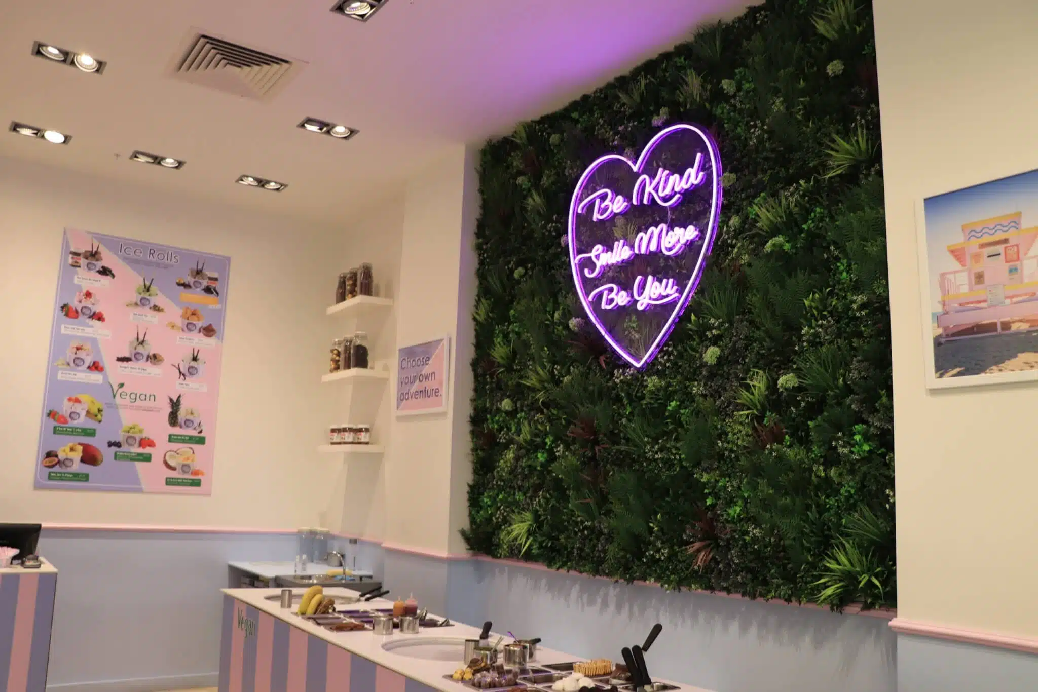 A modern cafe interior with a plant-covered wall featuring a heart-shaped neon sign that reads, Be Kind, Smile More, Be You. The counter displays dessert ingredients, and the walls have posters featuring vegan options.