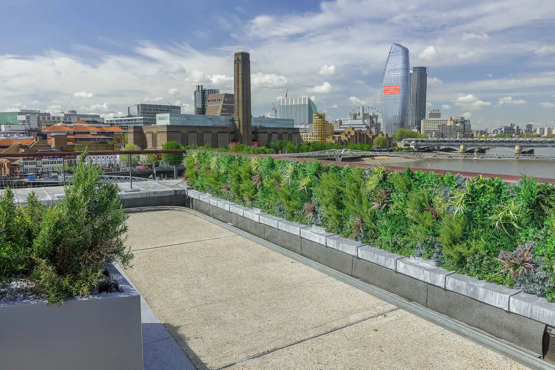 View of a rooftop garden with lush green plants overlooking a cityscape. The skyline features modern and historic buildings, including a tall, uniquely-shaped glass structure. A river is visible beside the city under a partly cloudy sky.