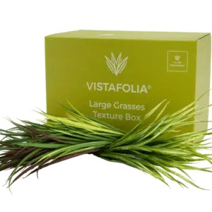 A green Vistafolia package, labeled Large Grasses Texture Box, is displayed with an array of artificial grass blades in shades of green and burgundy arranged before it.