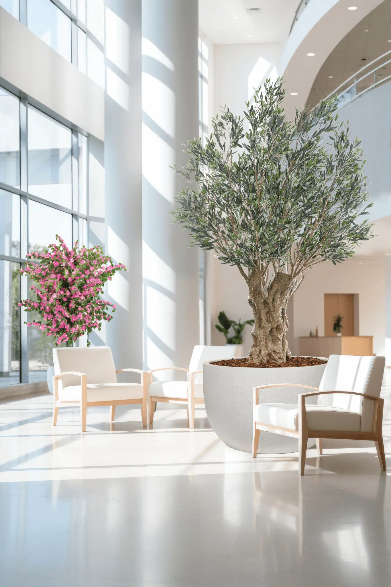 A modern, bright lobby features large windows for abundant sunlight, white armchairs with wood framing, and two Artificial Trees & Bushes: one with lush green leaves and another with pink flowers, both casting soft shadows on the floor.