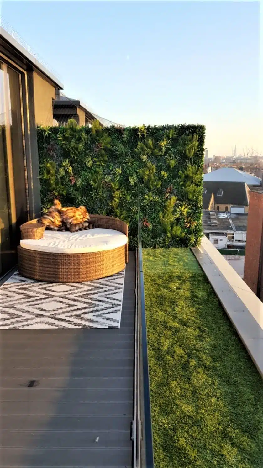 A rooftop terrace with a wicker round daybed featuring colorful cushions. The area includes a decorative green wall and a grass strip beside a gray deck, with a patterned rug. The skyline and rooftops are visible in the background at sunset.