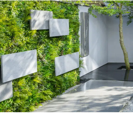 A modern outdoor area features living wall panels filled with lush greenery and complemented by rectangular stone surfaces. A smooth concrete path guides visitors to a small courtyard, where a single tree grows from the floor. Natural light brightens the entire space.