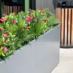 A large gray planter filled with lush green foliage and vibrant red flowers is situated on a paved outdoor area. A wooden and metal trash can is visible in the background.