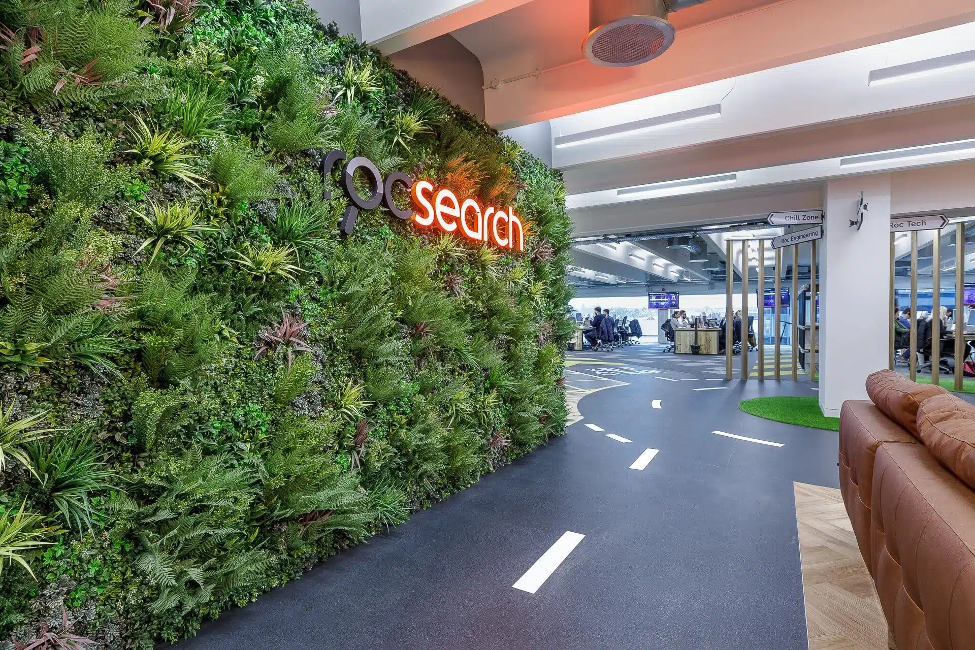 Office interior with a vertical green wall displaying search in red and white lettering. A road-themed carpet leads to an open seating area and meeting space. Modern design with natural and tech elements.