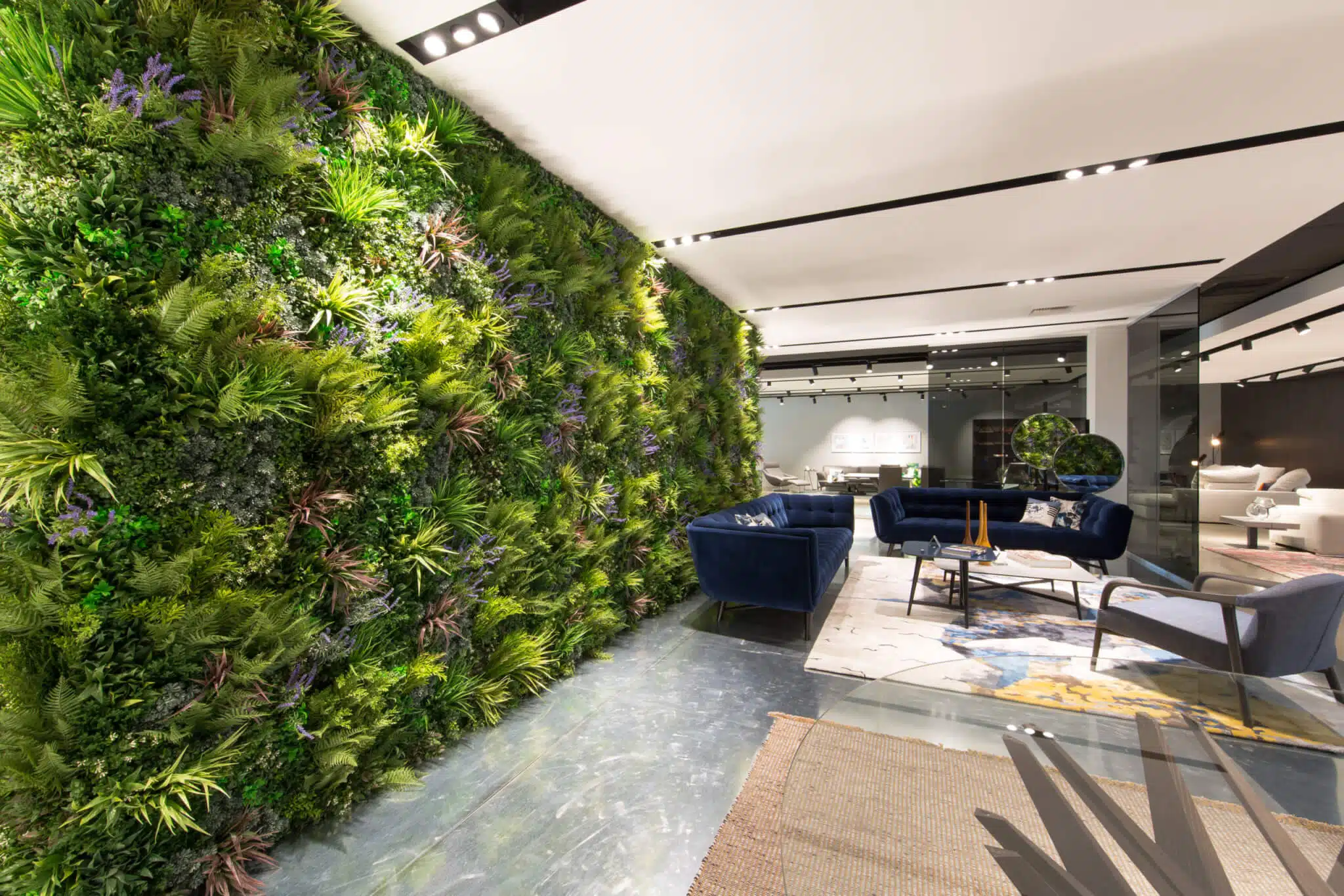 A modern indoor space has a vertical green plant wall on the left, blue sofas in the seating area, and a Soft Lavender Colour Box adding charm to the sleek decor. This stylish arrangement is centered around a round coffee table on the right.