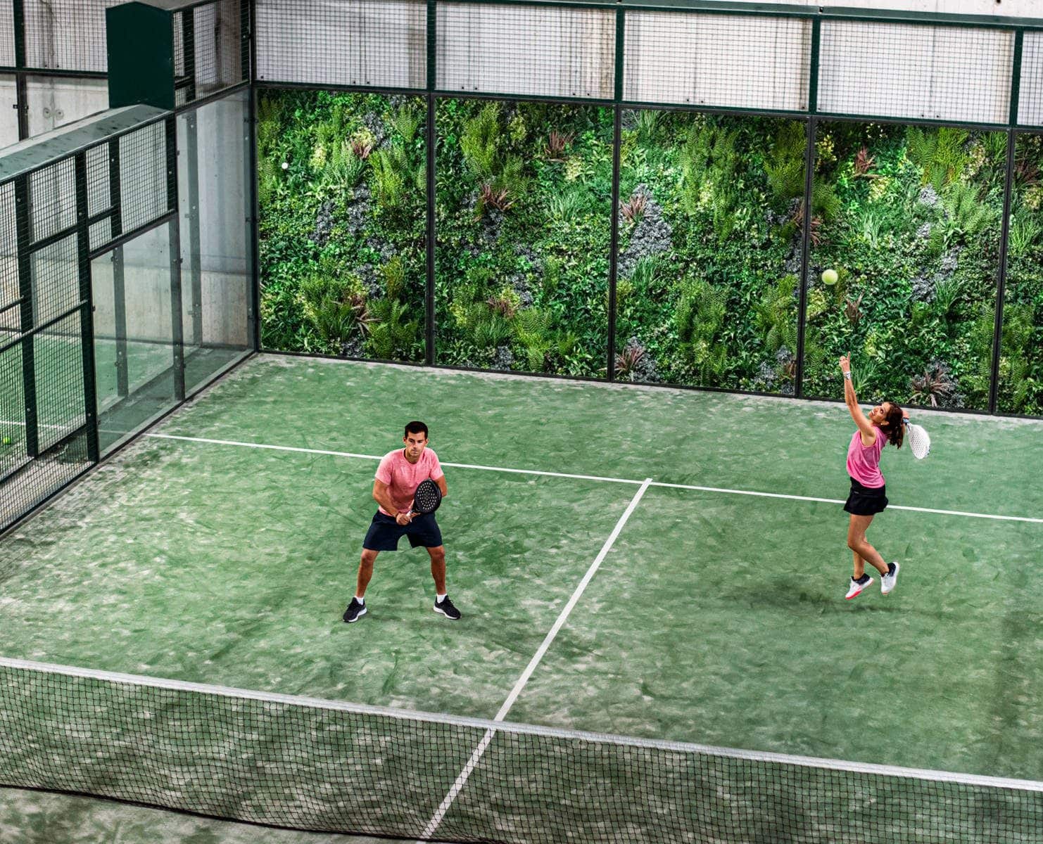 Two people playing padel in an indoor court with green walls. One person is preparing to hit the ball with a racket, while the other stands ready. Both are wearing pink tops and black shorts.