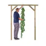 A man in a navy shirt and gray pants stands on a stool, hanging the Extendable Artificial Trailing Foliage on a wooden frame against a plain white background.