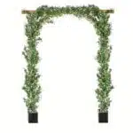 The Extendable Artificial Trailing Foliage creates an elegant archway with lush greenery and delicate white flowers, supported by black planters. Crafted from wooden beams, this charming structure stands gracefully against a plain white background.