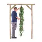 A man in a navy shirt and gray pants decorates a wooden arch with the Extendable Artificial Trailing Foliage. He stands on a stool, reaching up to adjust the greenery.
