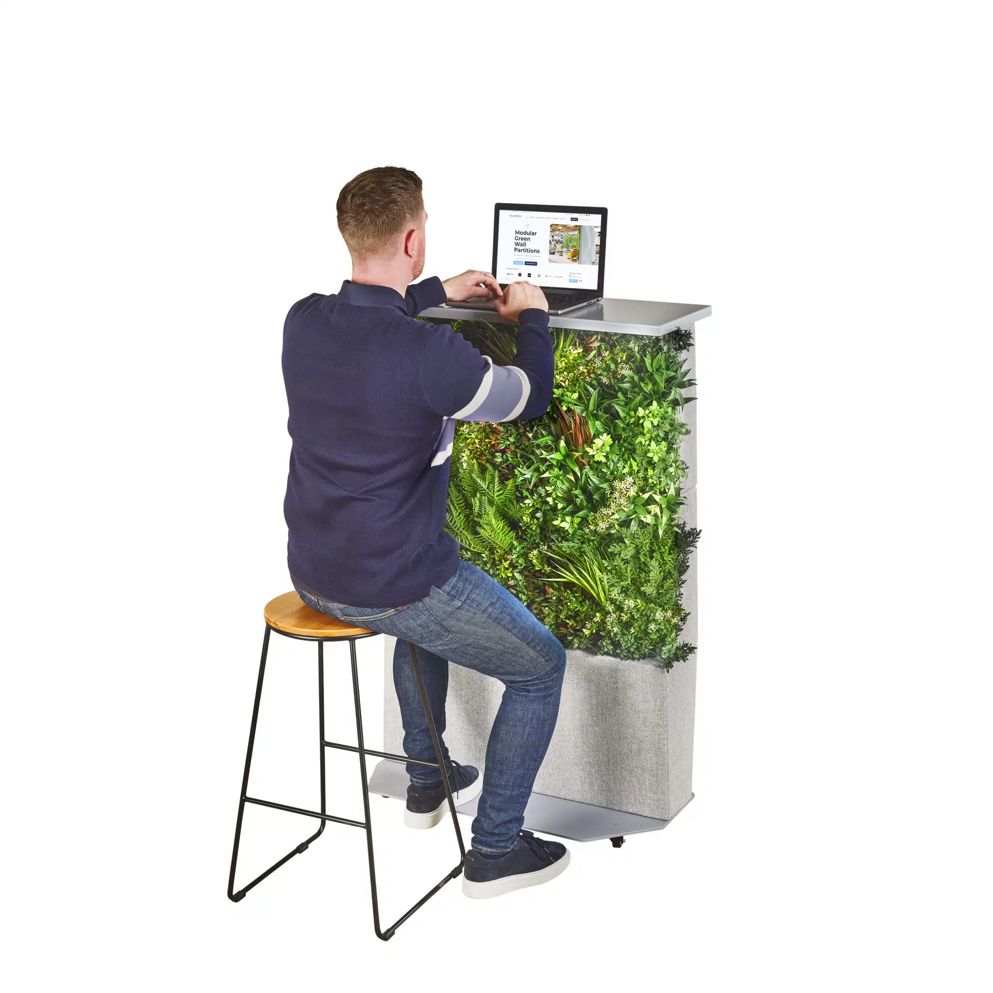 Someone in a dark sweater sits on a high stool working at a standing desk, featuring Modular Green Wall Partitions filled with lush plants. The background is white.