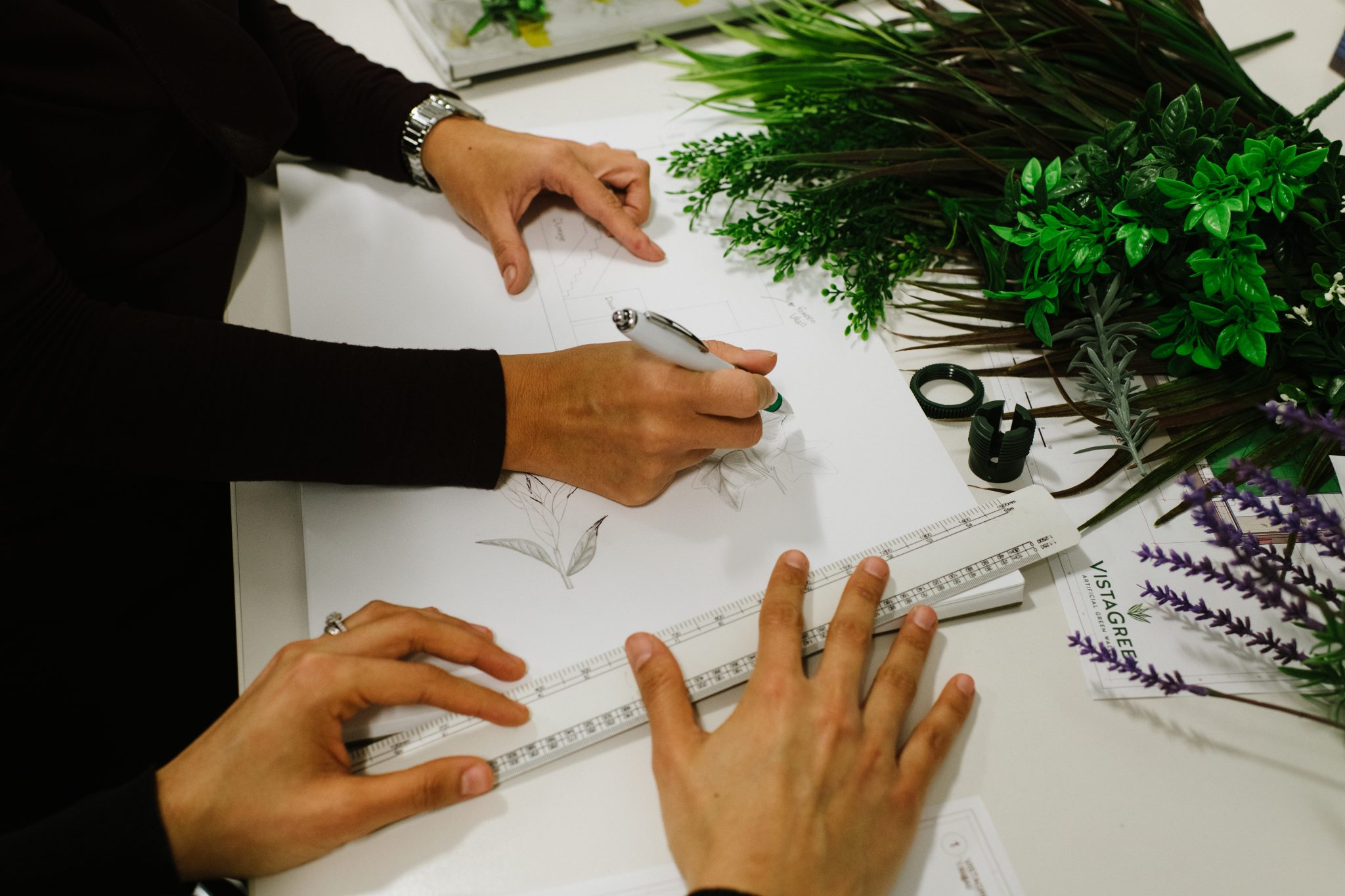 Two people are working on botanical sketches at a table. One uses a pen to draw, while the other holds a ruler on the paper. Various green and purple artificial plants are scattered on the table nearby.
