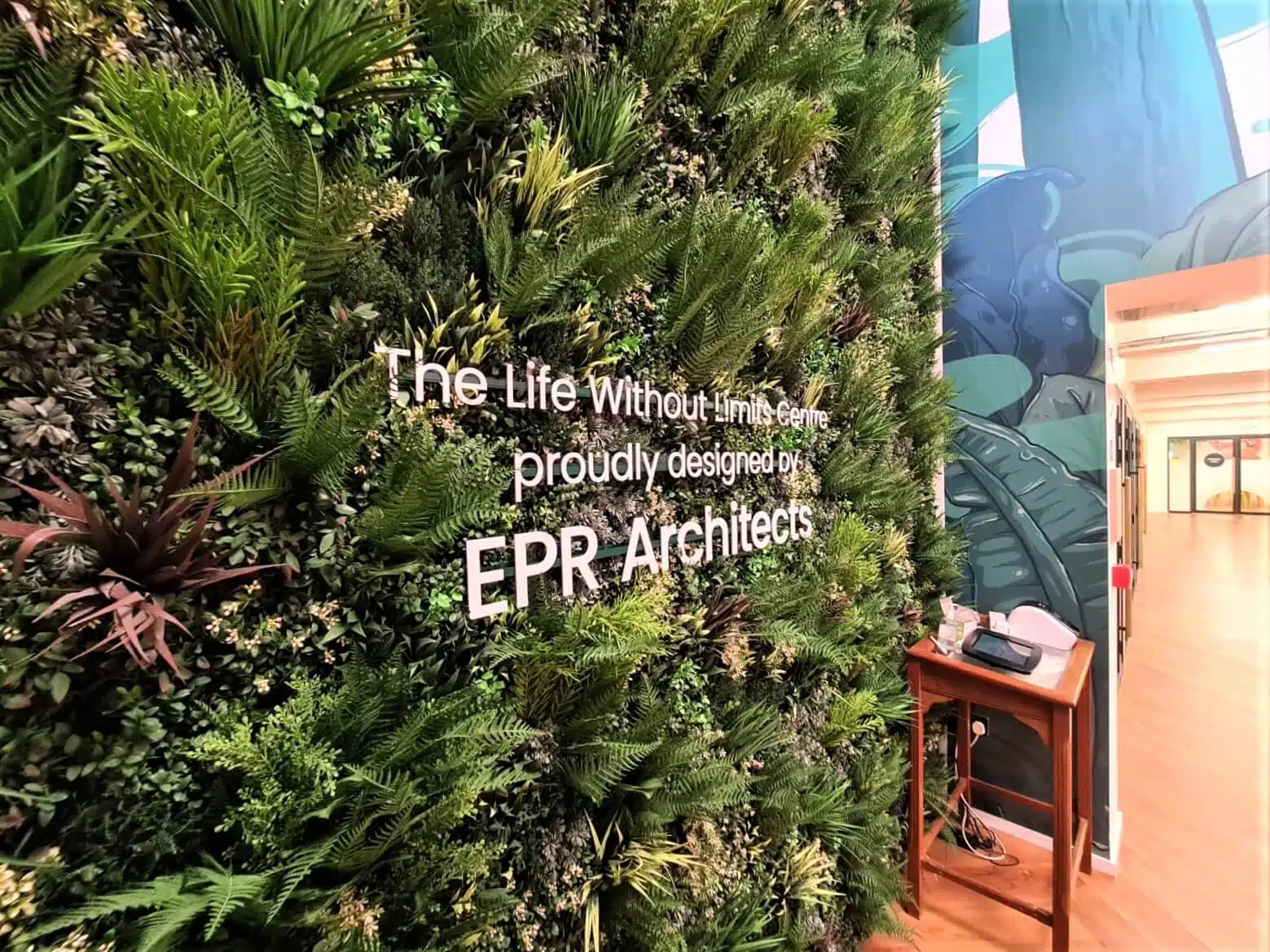 A vertical plant wall with the text The Life Without Limits Centre, proudly designed by EPR Architects is displayed. The area is bright with a wooden floor, and a small table with a device is visible at the side.
